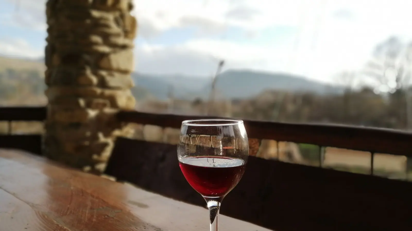 A single glass of red wine on a wooden ledge, with an open valley and hills out of focus beyond it.