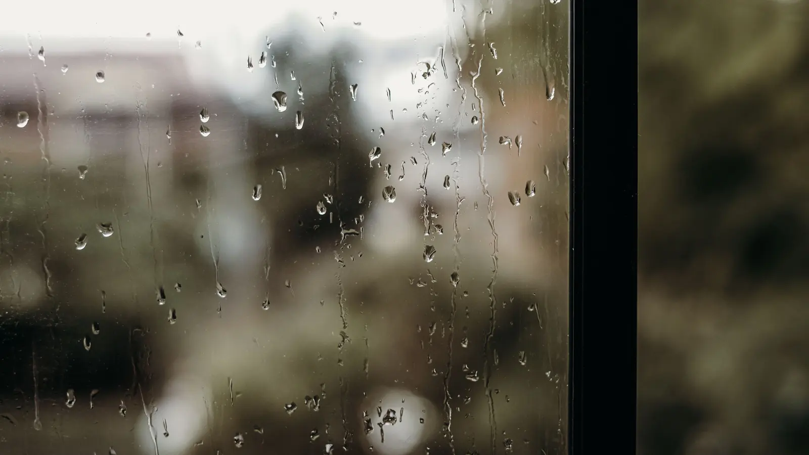 Raindrops running down a window pane, with a blurred view of houses and trees outside.