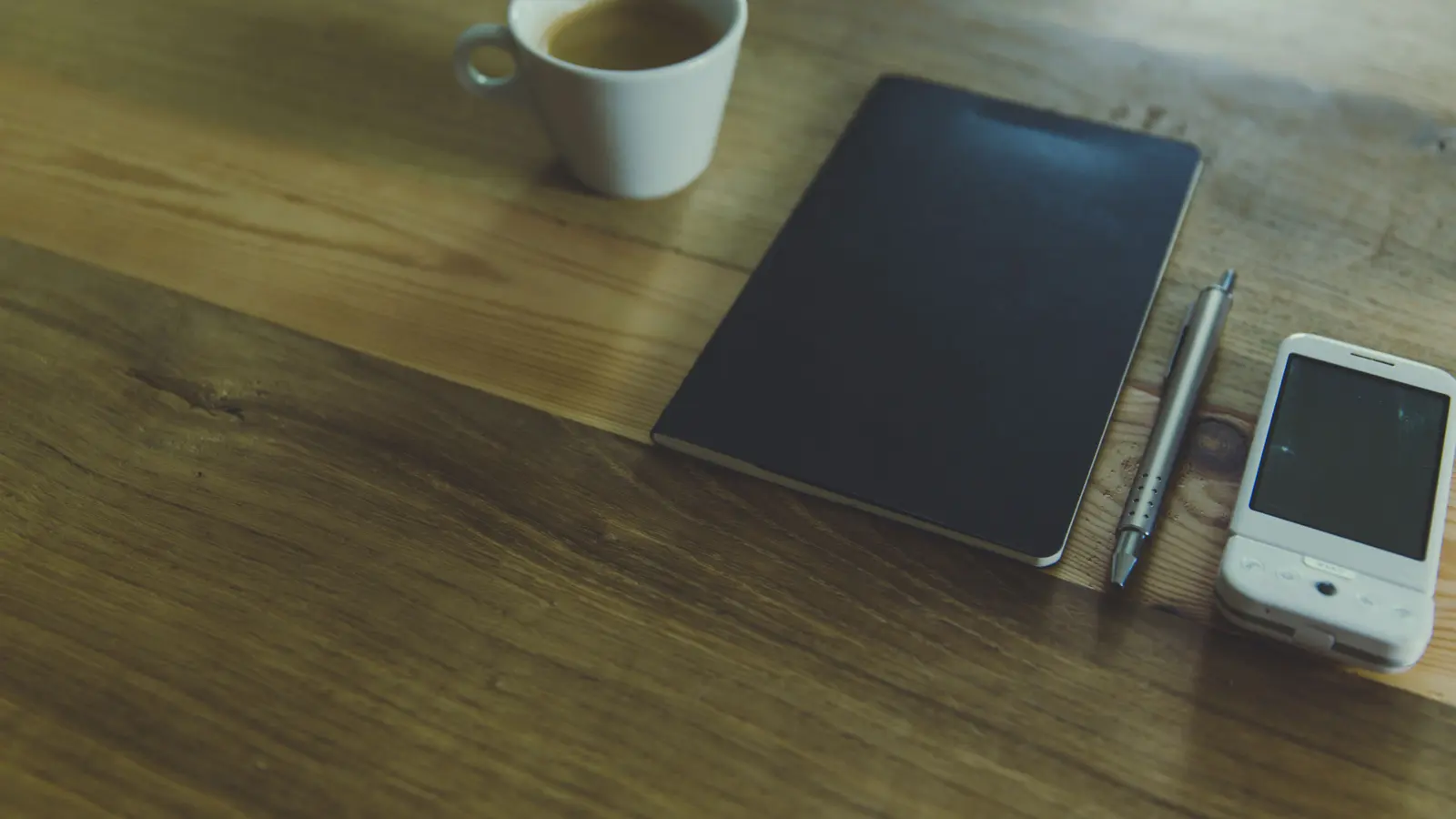 A white mug of coffee, a notebook and a pen arranged on a wooden desk, photographed from above in flat daylight.
