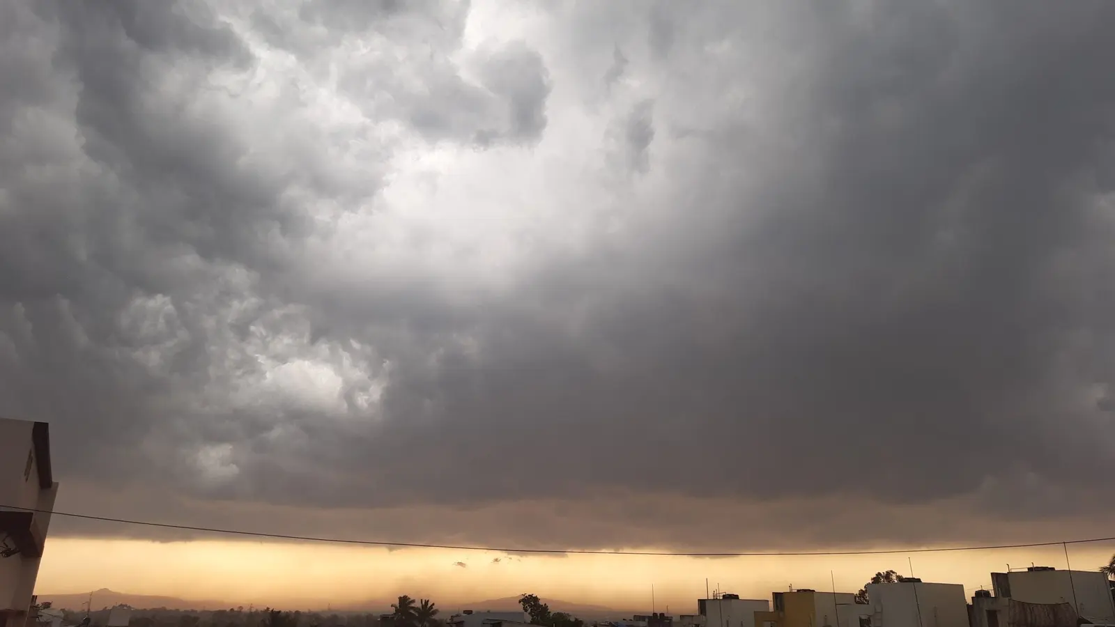 Thick dark storm cloud filling the sky above a town's rooftops, with a narrow band of amber light along the horizon beneath it.