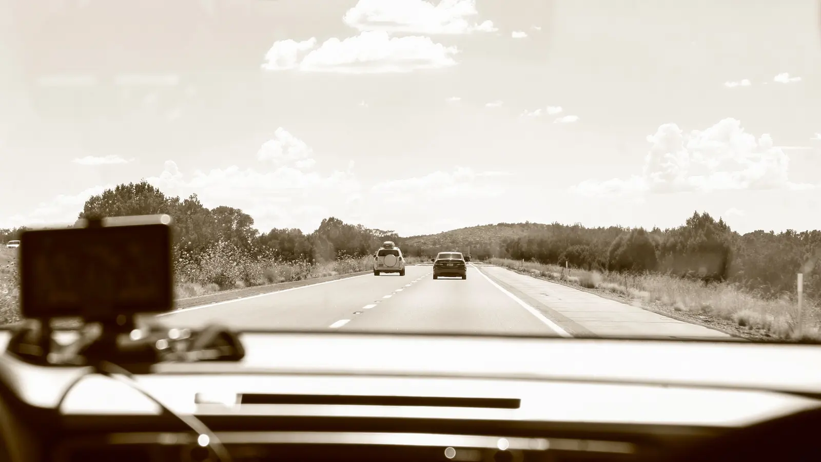 The view through a car windscreen along a straight open road, with two cars ahead and countryside on both sides.