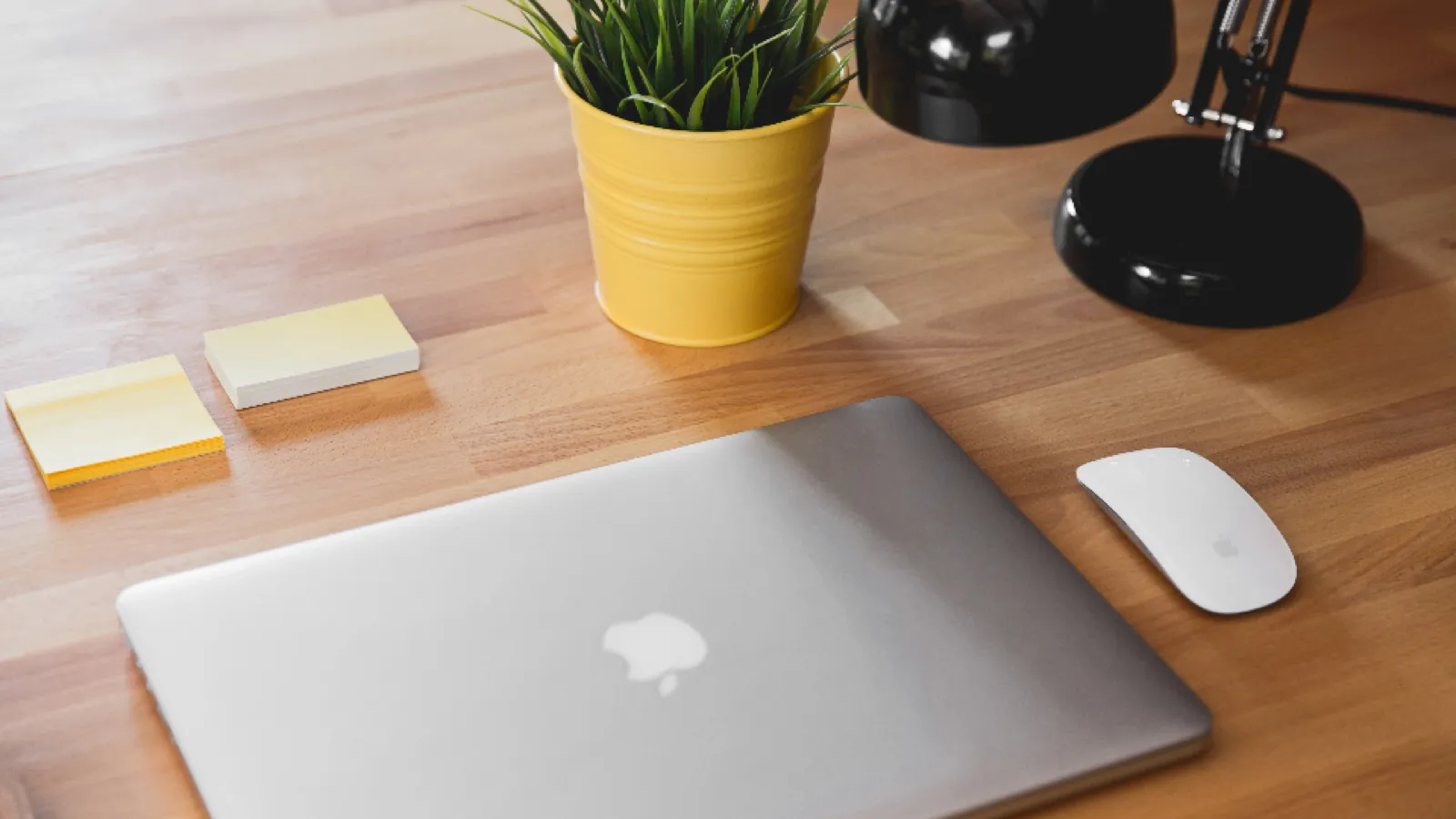 A wooden desk seen from above: a closed laptop, a white mouse, two pads of sticky notes, a plant in a yellow pot, and an angled black desk lamp.