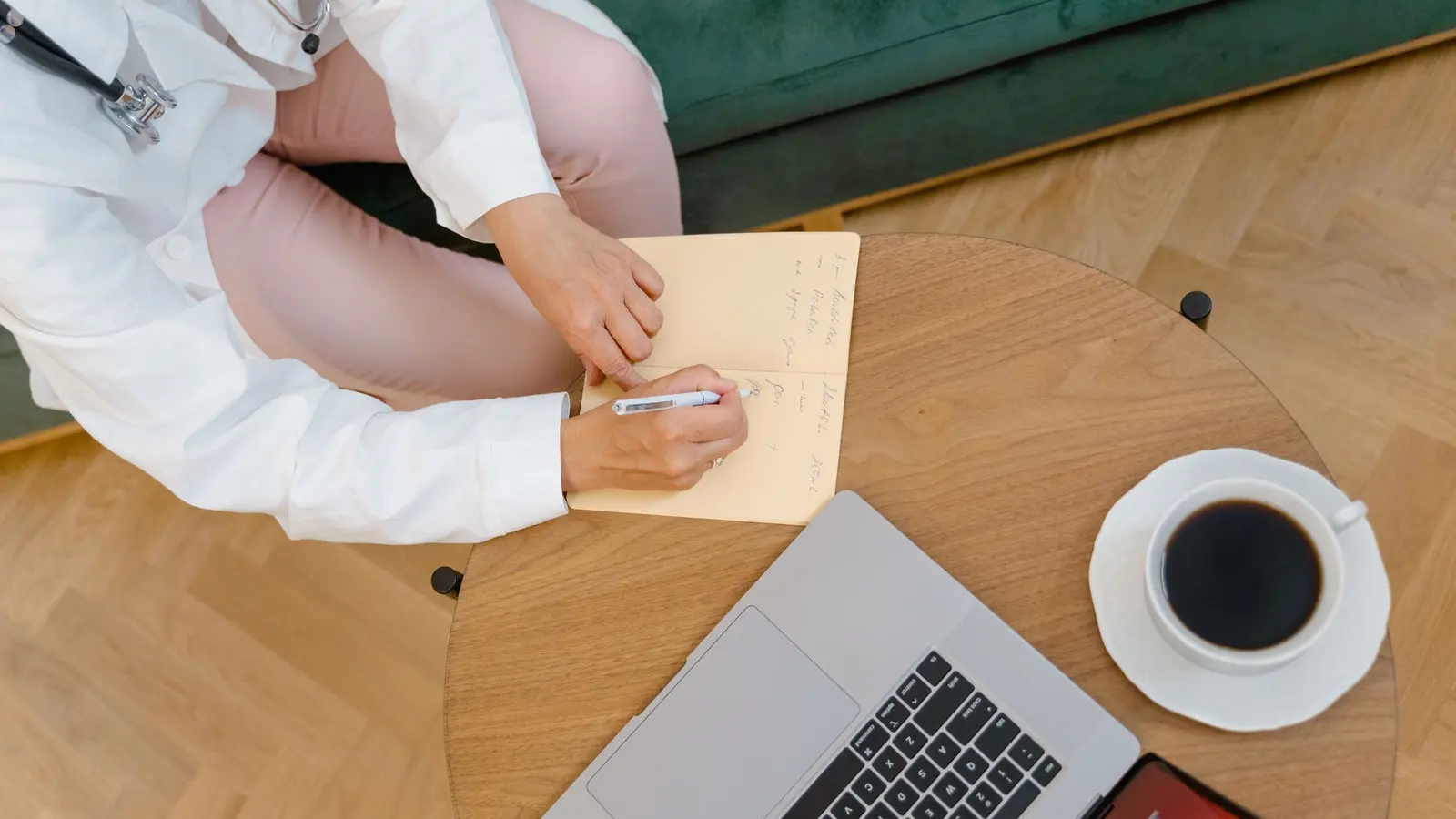 An overhead view of a clinician in a white coat and stethoscope writing in a notebook at a round wooden table, a laptop and a cup of black coffee beside it.