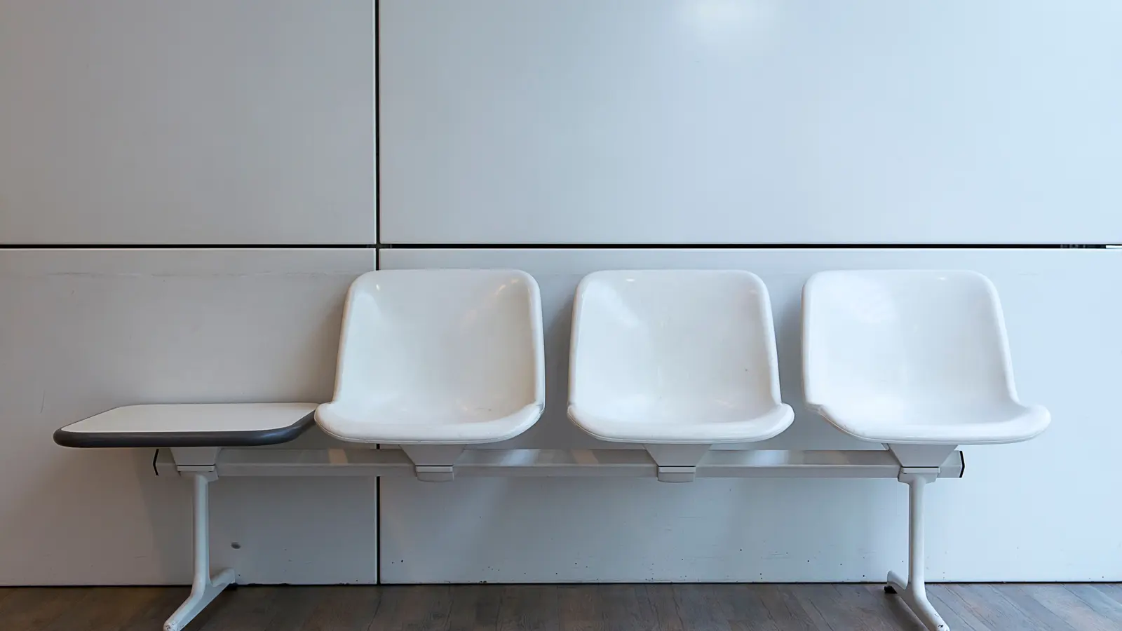 A row of moulded white waiting-room chairs against a plain white wall, with a small flat side table at one end.