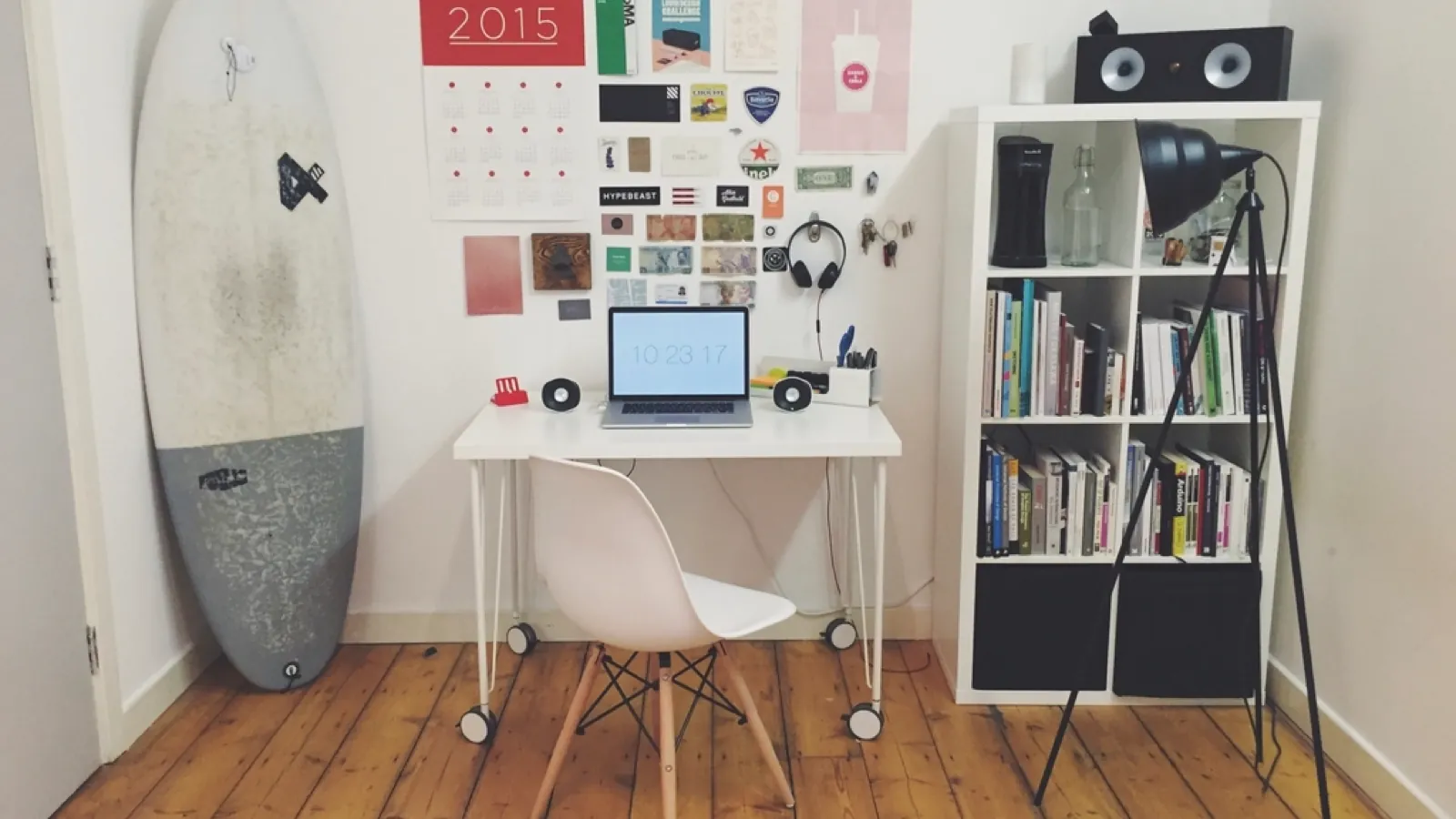 A teenager's room: a surfboard against the wall, a pinboard covered in photos and stickers above a small white desk with an open laptop, and a bookshelf beside it.