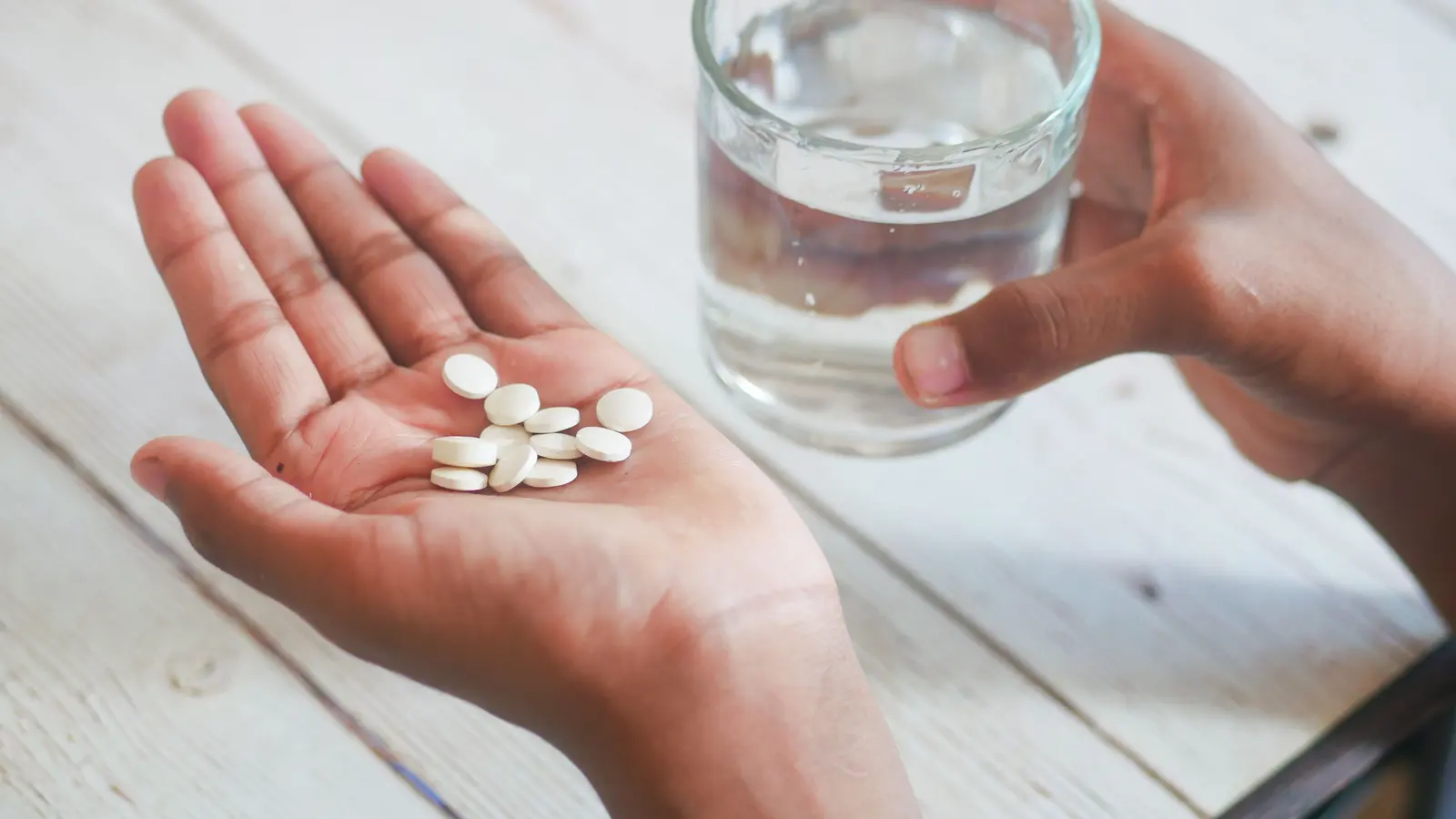 An open palm holding several small white tablets, with the other hand holding a glass of water above a pale whitewashed wooden table.