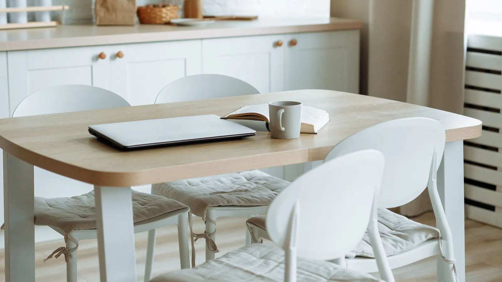 A closed laptop, a grey mug and an open notebook on a pale wooden kitchen table, with white chairs pushed in and a bright kitchen behind.