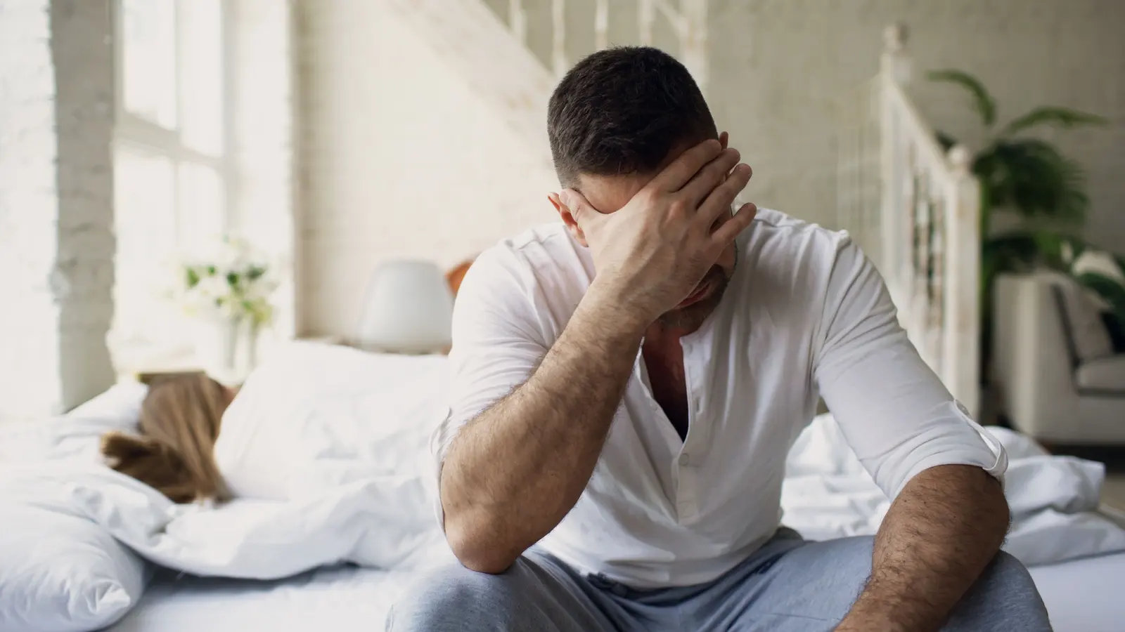 A man sits on the edge of the bed in a bright morning bedroom, one hand covering his eyes, while someone sleeps on behind him.