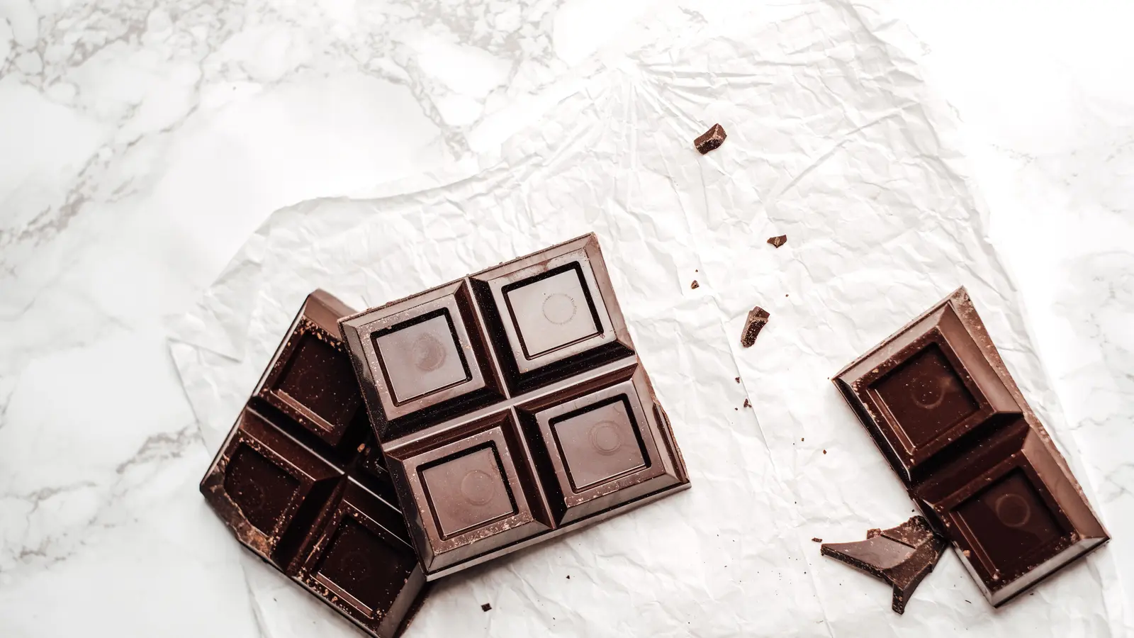 Des carrés de chocolat noir brisés sur du papier blanc froissé, au-dessus d'une surface en marbre pâle, photographiés du dessus.