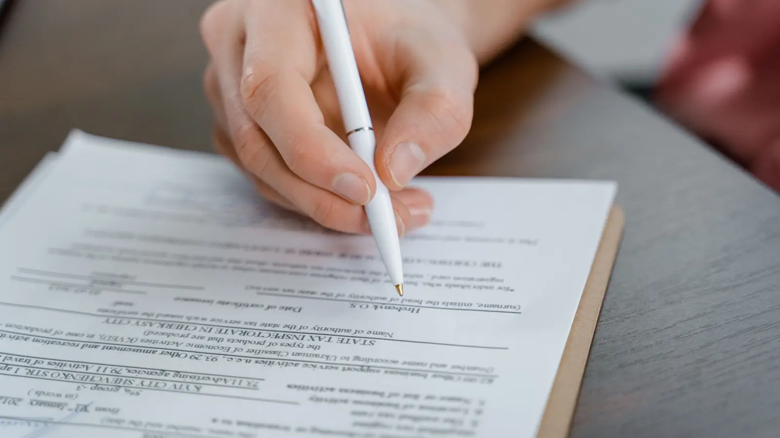 A hand holding a white pen and filling in a printed form resting on a clipboard at a desk.