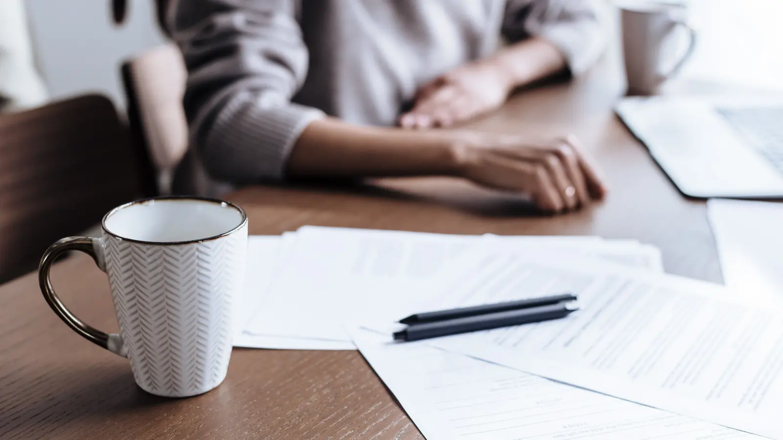 A patterned mug beside loose printed papers and a black pen on a dark wooden table, a person's hands resting out of focus behind them.