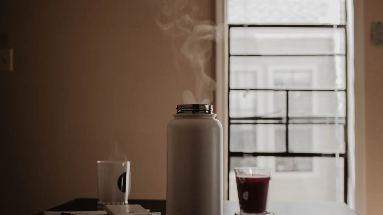 A steaming mug, a small thermos and an open notebook resting on a wooden table in warm window light.