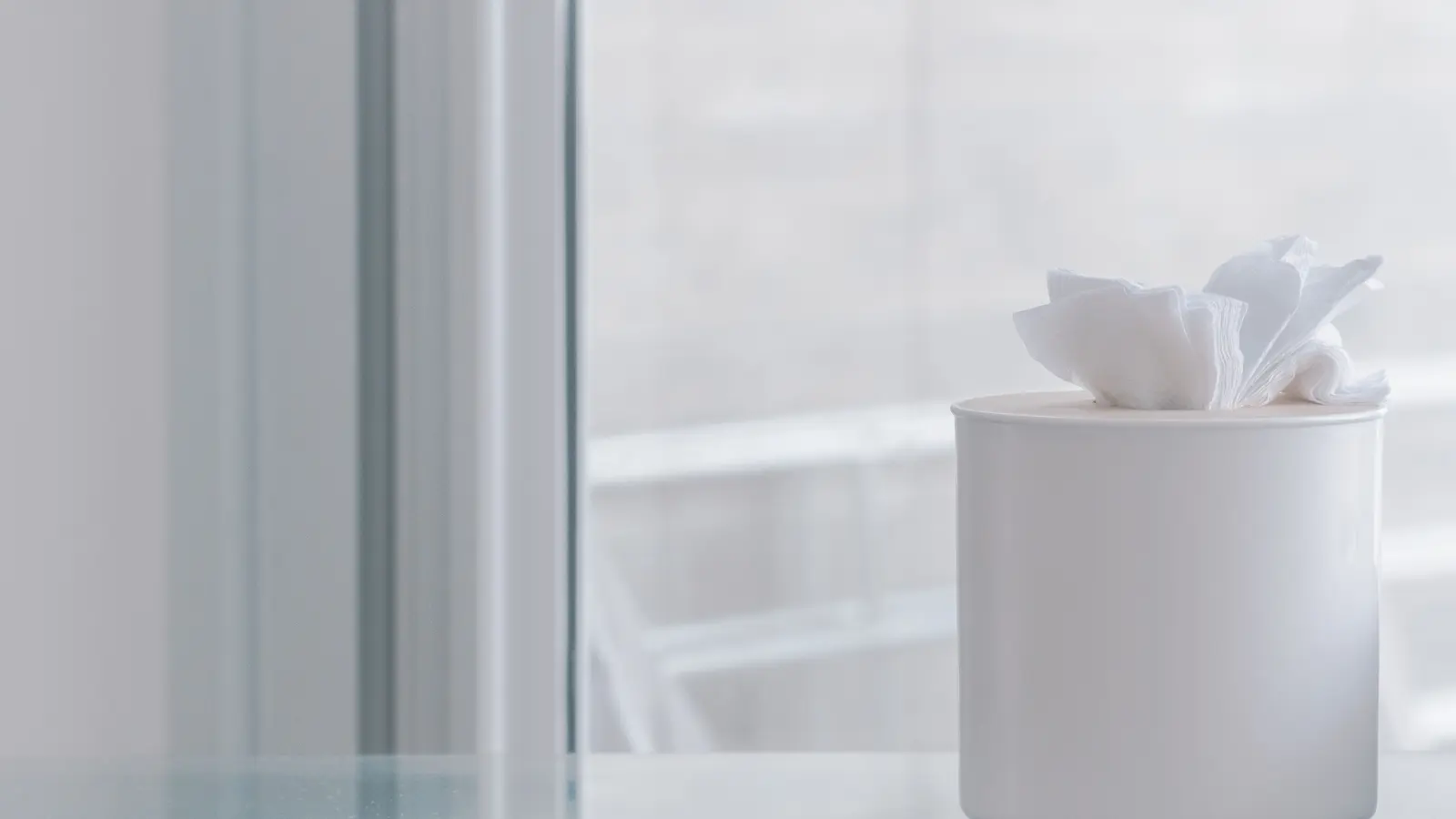 A plain white tissue holder on a glass table beside a bright window, the rest of the frame empty