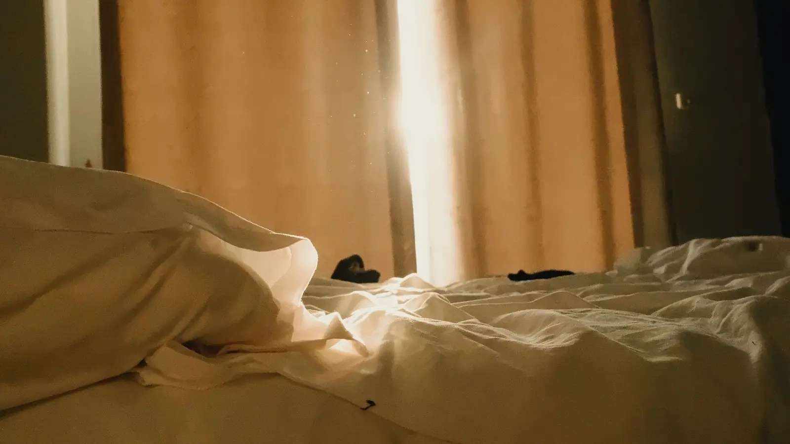 Warm morning sunlight streams through sheer curtains onto a cozy, unmade bed.