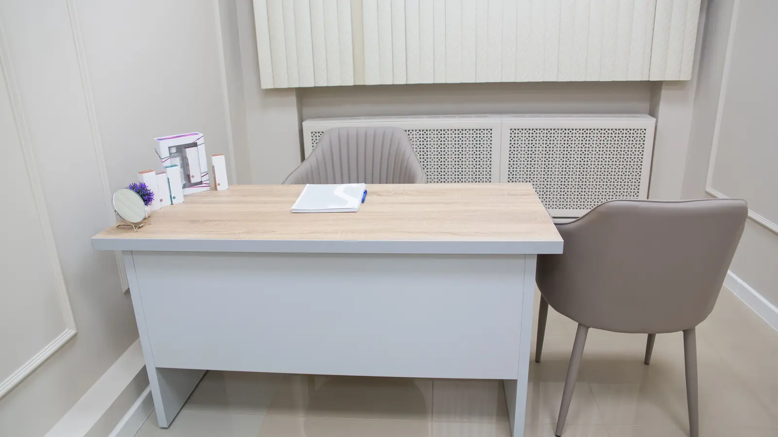 A quiet consulting room with a pale wooden desk, a chair on each side of it, and a notepad and pen set out on the surface.