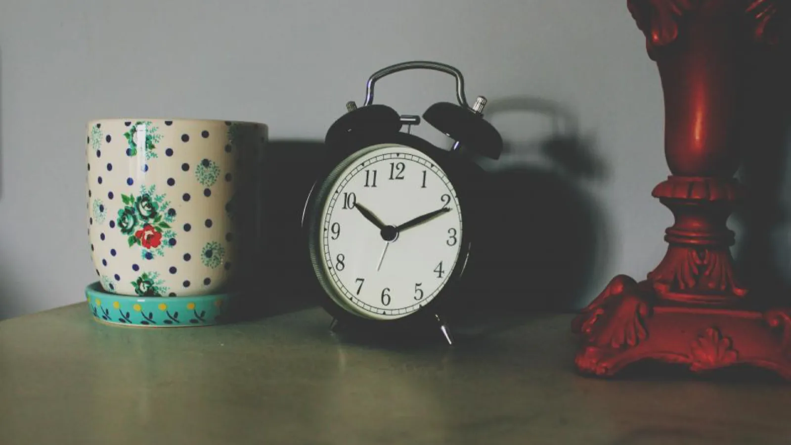 Objects on a bedside surface in dim light: a floral mug, a black twin-bell alarm clock, and the red base of a lamp.