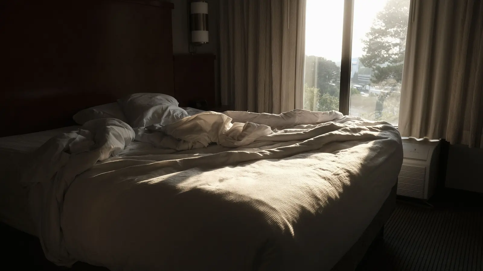 An unmade bed in a quiet bedroom with morning sunlight falling across the duvet from a window.