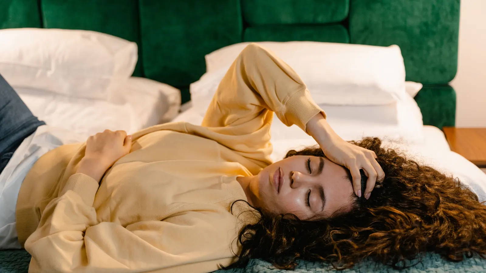 A woman lying on a bed with her eyes closed and one hand resting against her forehead, wearing a pale yellow sweatshirt against white pillows.